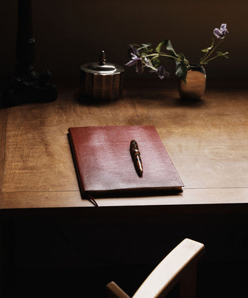 The Red Notebook with a lizard-embossed cover and a pen rests on a wooden desk near a small vase of purple flowers and a decorative container.