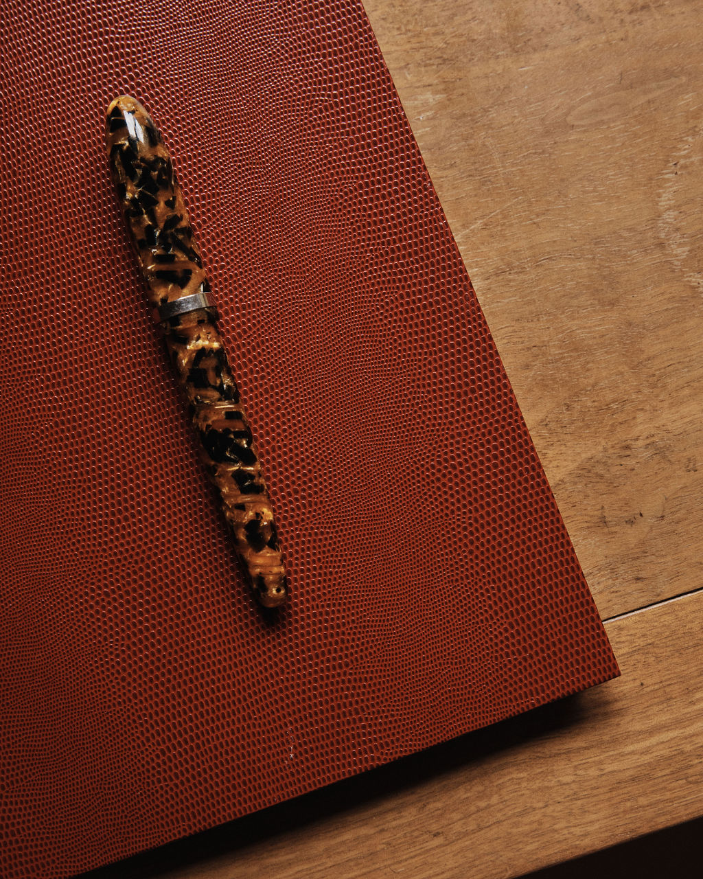 A marbled brown and black fountain pen rests on a textured, luxury Red Notebook placed on a wooden surface.