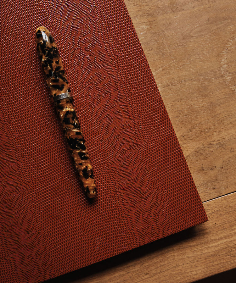 A marbled brown and black fountain pen rests on a textured, luxury Red Notebook placed on a wooden surface.