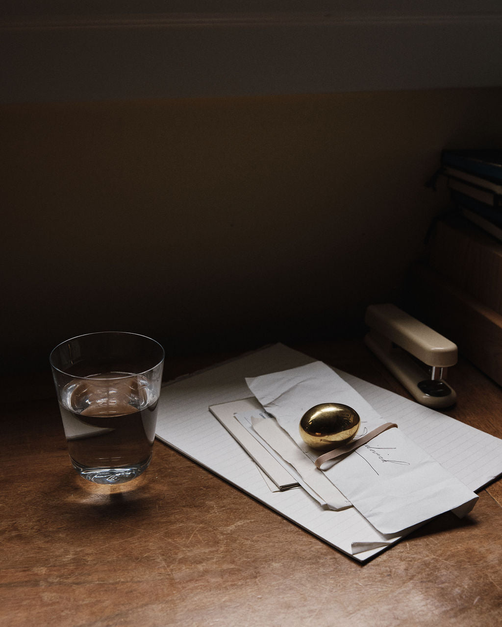 A glass of water, a golden spoon, some papers, and the Egg Paperweight—a sculptural brass accessory—are arranged on a wooden desk under soft lighting.