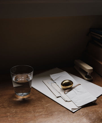 A glass of water, a golden spoon, some papers, and the Egg Paperweight—a sculptural brass accessory—are arranged on a wooden desk under soft lighting.