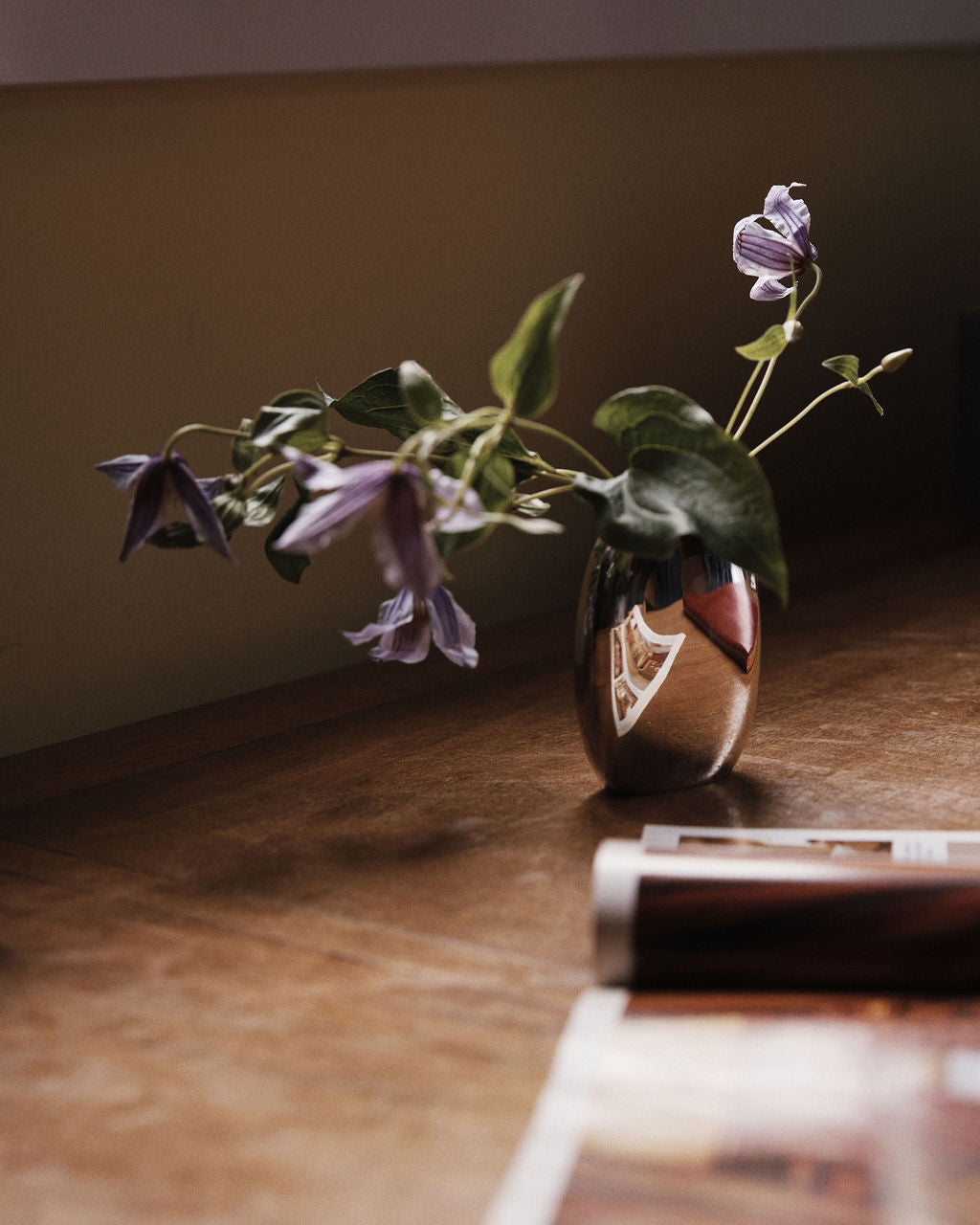 The Vera Oval Budvase, featuring a pewter finish, holds purple flowers as it rests on a wooden surface beside a magazine, its soft reflection visible on the vase.