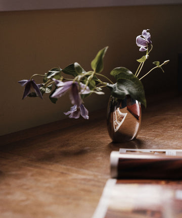 The Vera Oval Budvase, featuring a pewter finish, holds purple flowers as it rests on a wooden surface beside a magazine, its soft reflection visible on the vase.