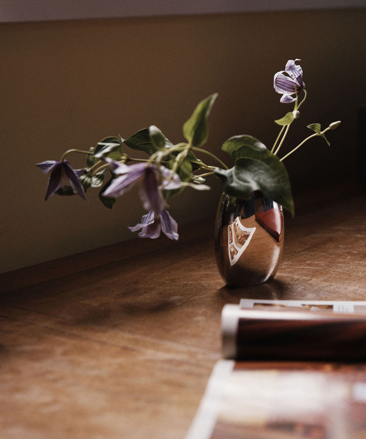 The Vera Oval Budvase, featuring a pewter finish, holds purple flowers as it rests on a wooden surface beside a magazine, its soft reflection visible on the vase.