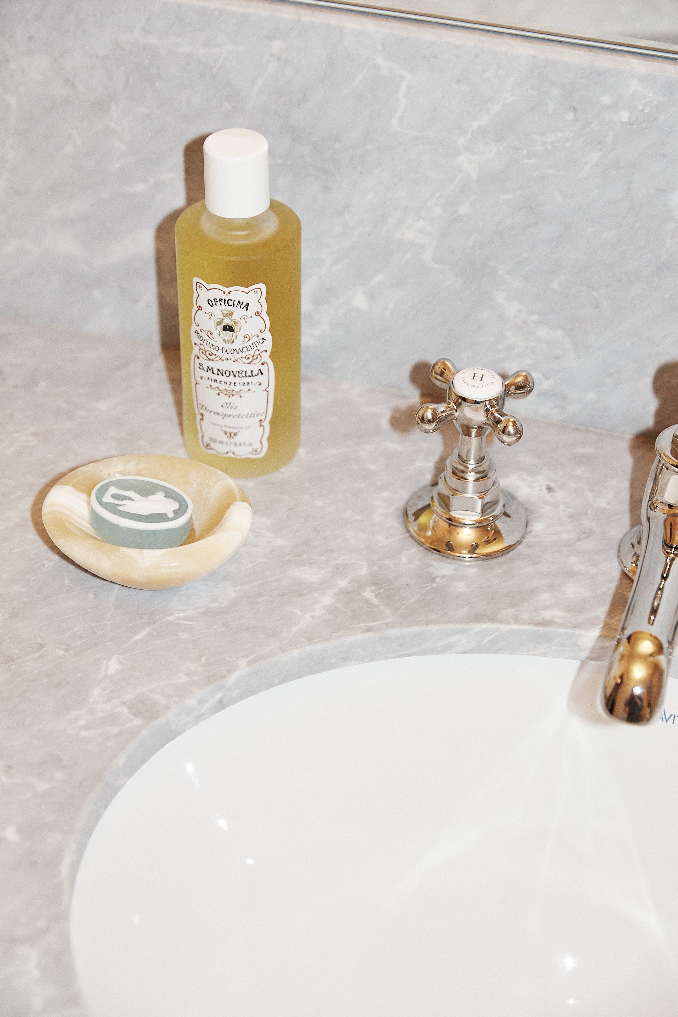 A bottle of yellow liquid, a decorative green soap in a dish, and a silver faucet sit on a gray marble bathroom countertop beside a white sink, complemented by the Akila Catchall, which adds a unique decorative accent to the space.