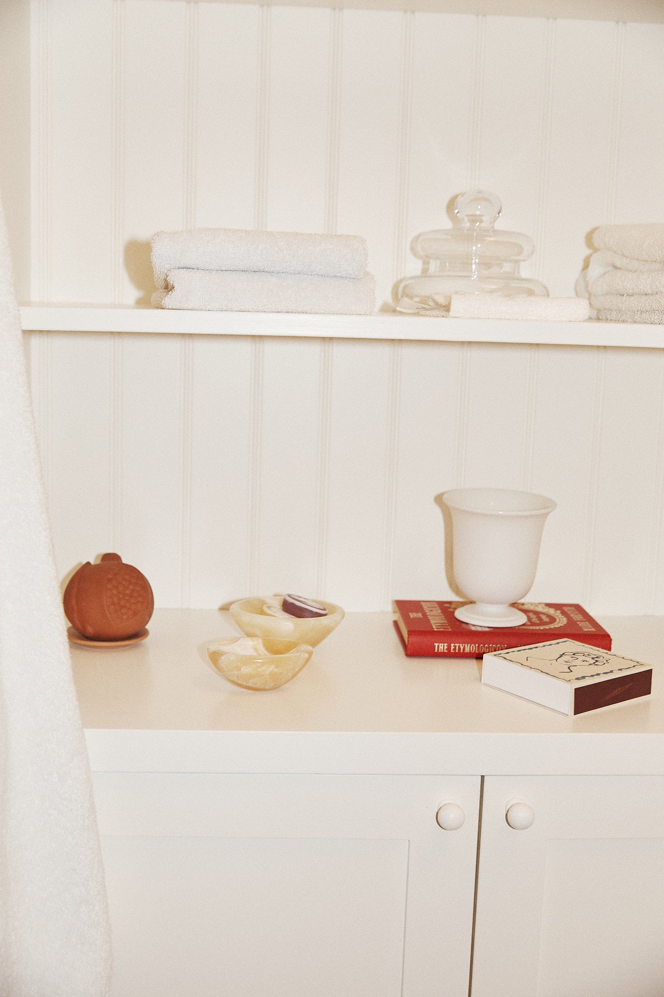 White cabinet with shelves holding folded towels, a glass jar, a red book, a white vase, decorative accents like the Akila Catchall and a decorative pumpkin, plus a dish with soap and a box of matches against a white wall.