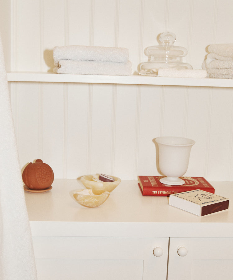 White cabinet with shelves holding folded towels, a glass jar, a red book, a white vase, decorative accents like the Akila Catchall and a decorative pumpkin, plus a dish with soap and a box of matches against a white wall.