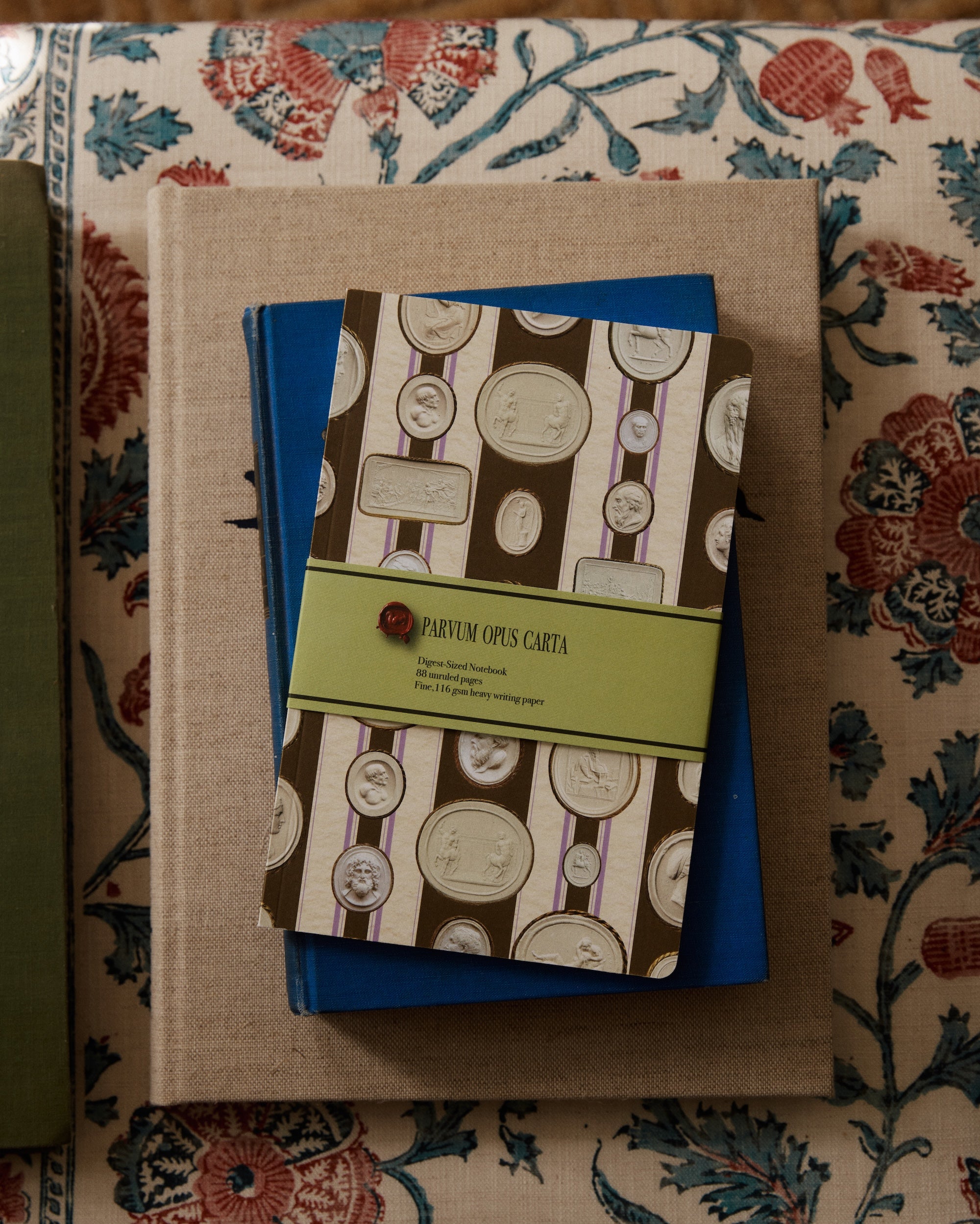 An Intaglio Notebook with a coin pattern and green label rests on blue and beige books atop patterned fabric, all with acid-free writing paper.