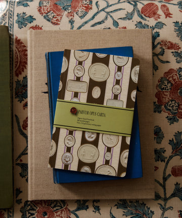 An Intaglio Notebook with a coin pattern and green label rests on blue and beige books atop patterned fabric, all with acid-free writing paper.