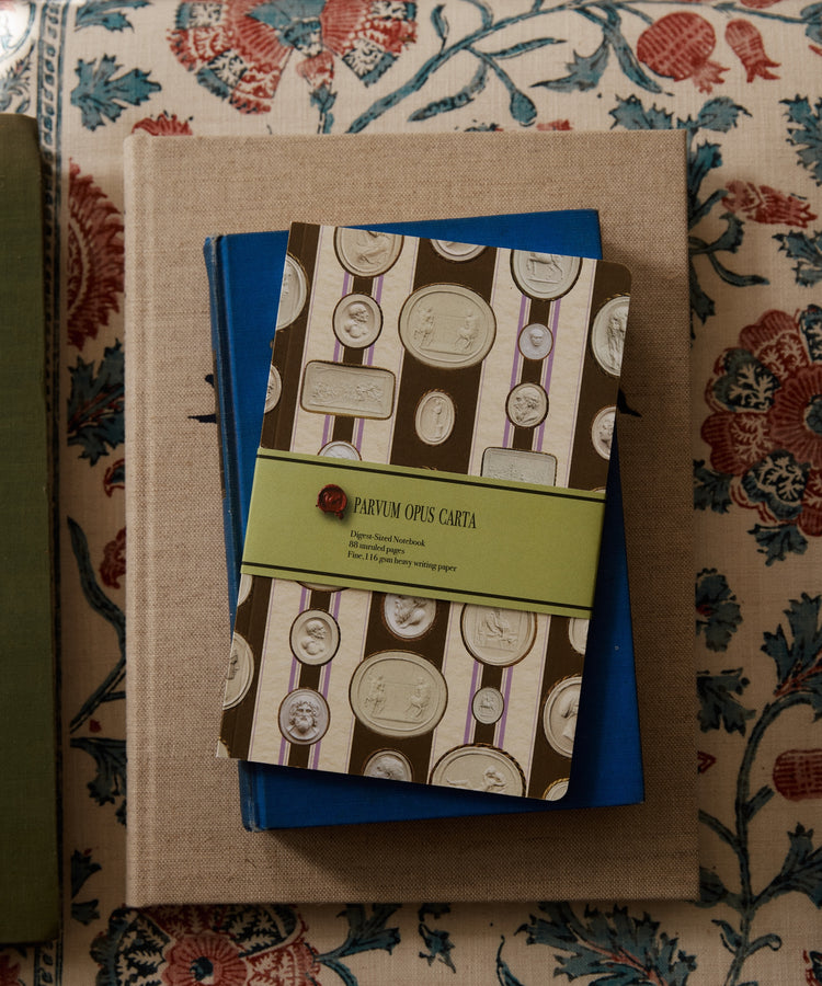 An Intaglio Notebook with a coin pattern and green label rests on blue and beige books atop patterned fabric, all with acid-free writing paper.