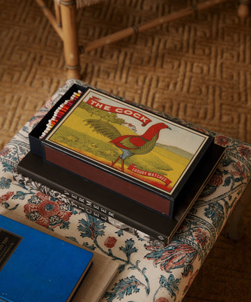 A box of Luxury Matches with a rooster design sits on a floral table, making a unique gift next to books.