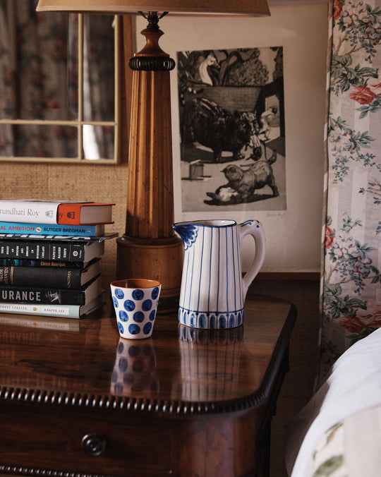 A wooden bedside table holds the Jorge Jug, a lamp, stacked books, and a blue polka dot cup; floral wallpaper and framed art decorate the background.