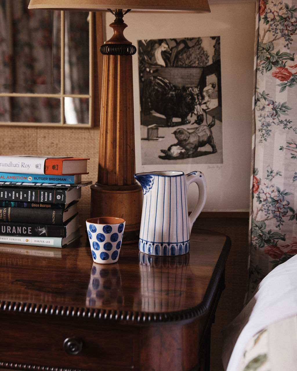 A wooden nightstand holds a stack of books, the Jorge Jug— a green-and-white striped ceramic pitcher resembling a Portuguese jug—a polka dot cup, and a lamp; floral wallpaper and framed art are in the background.