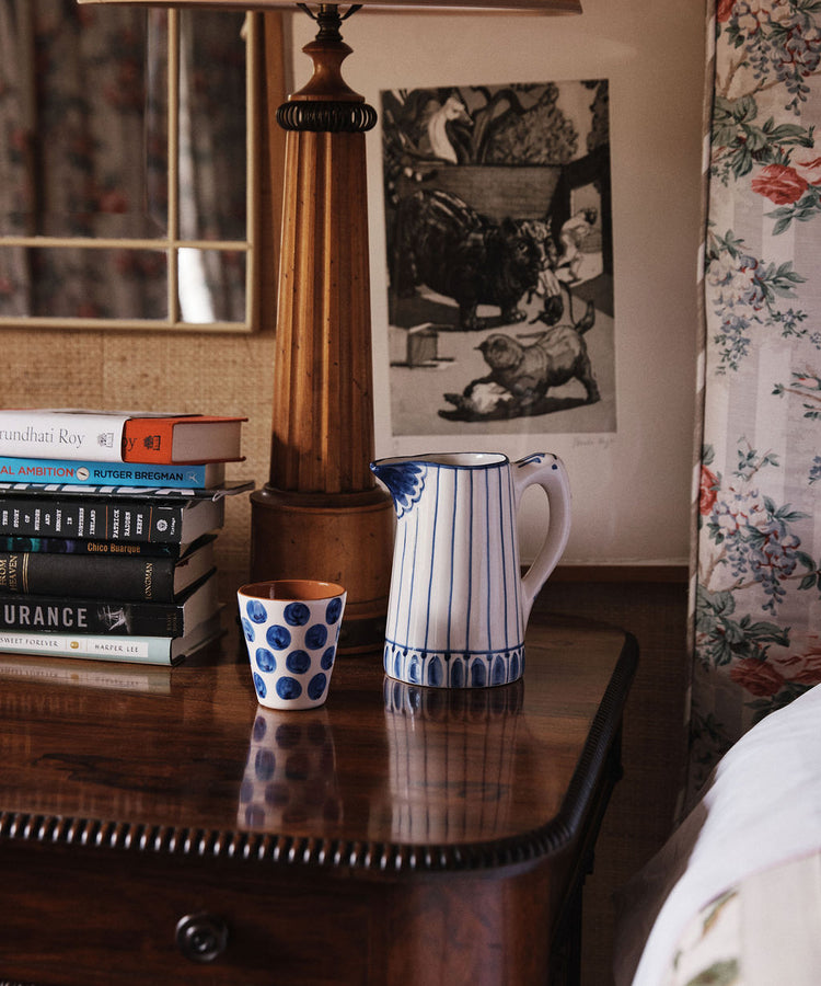A wooden nightstand holds a stack of books, the Jorge Jug— a green-and-white striped ceramic pitcher resembling a Portuguese jug—a polka dot cup, and a lamp; floral wallpaper and framed art are in the background.