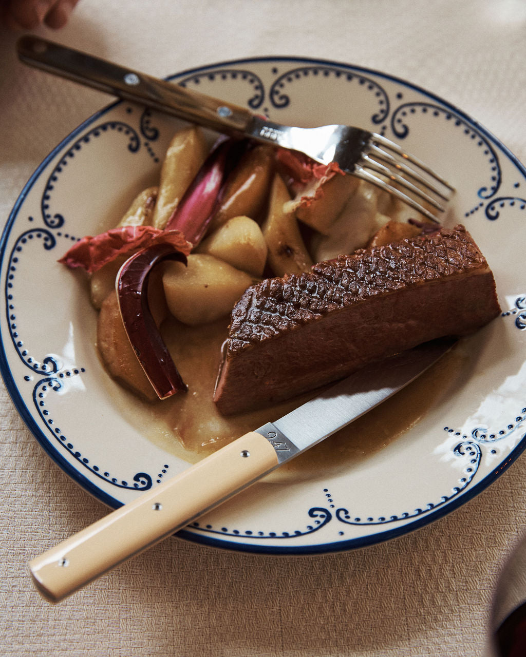 A plate with seared meat, roasted potatoes, and rhubarb is served with a handmade 9.47 Table Knife and fork on a decorative plate, showcasing French craftsmanship.