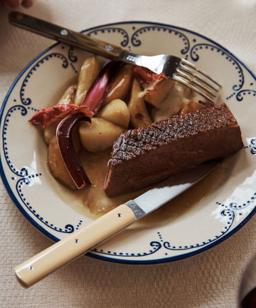 A plate with seared meat, roasted potatoes, and rhubarb is served with a handmade 9.47 Table Knife and fork on a decorative plate, showcasing French craftsmanship.