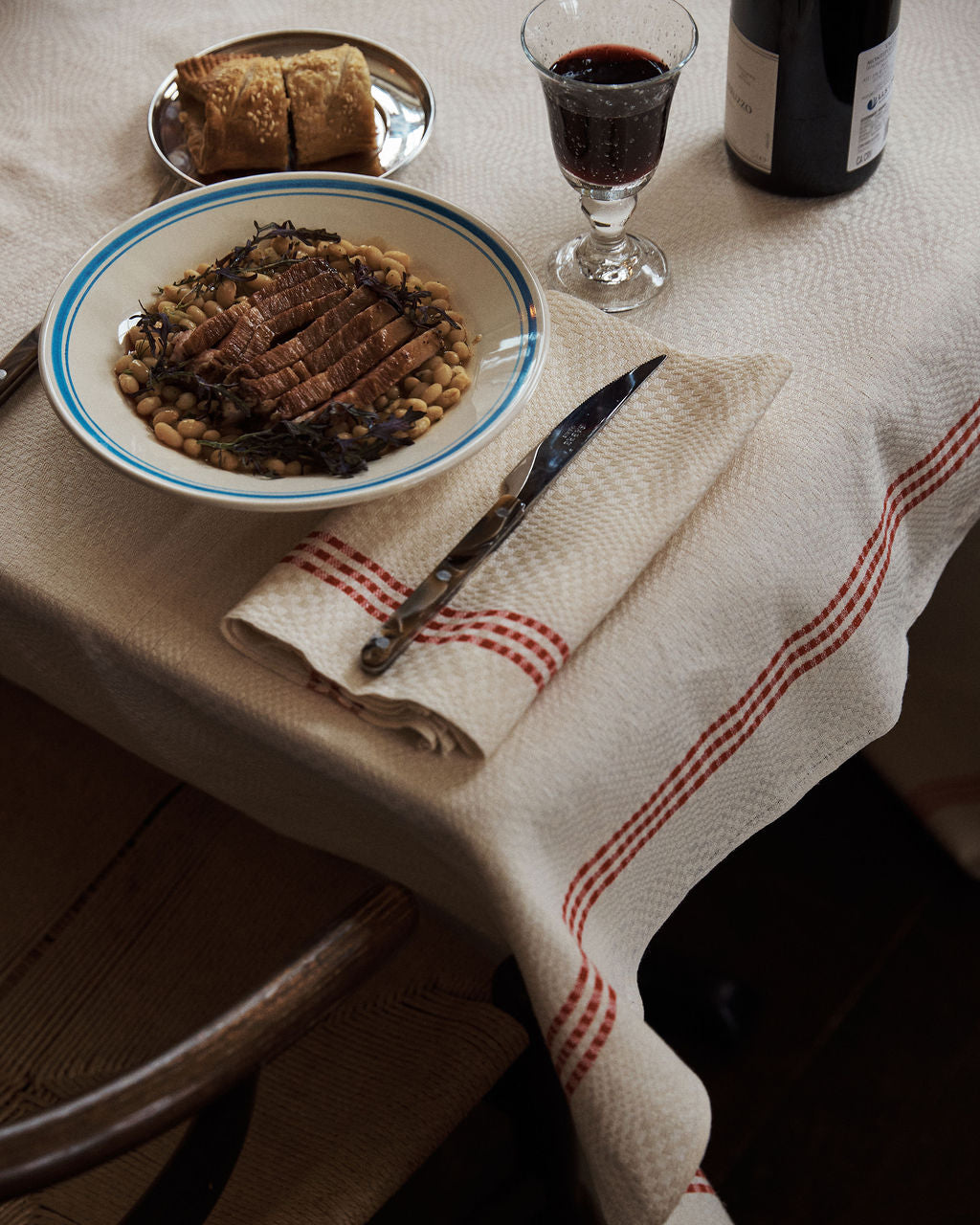 A table set with the Folk Dinner Plate, Jester, holds sliced meat on beans, accompanied by a glass of red wine, bread on a small plate, and cutlery resting on a folded napkin.
