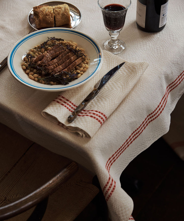 A table set with the Folk Dinner Plate, Jester, holds sliced meat on beans, accompanied by a glass of red wine, bread on a small plate, and cutlery resting on a folded napkin.