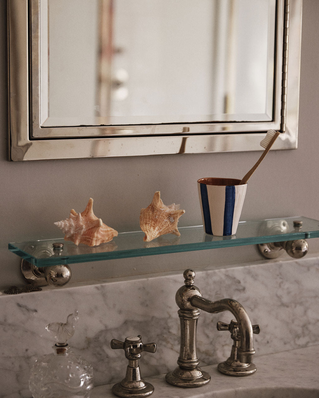 A bathroom sink with a marble countertop, glass shelf with two seashells, a Lula Cup, Set of 6, holding a toothbrush, and a rectangular mirror above.