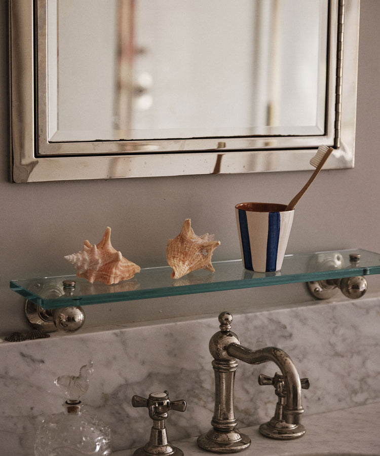 A bathroom sink with a marble countertop, glass shelf with two seashells, a Lula Cup, Set of 6, holding a toothbrush, and a rectangular mirror above.
