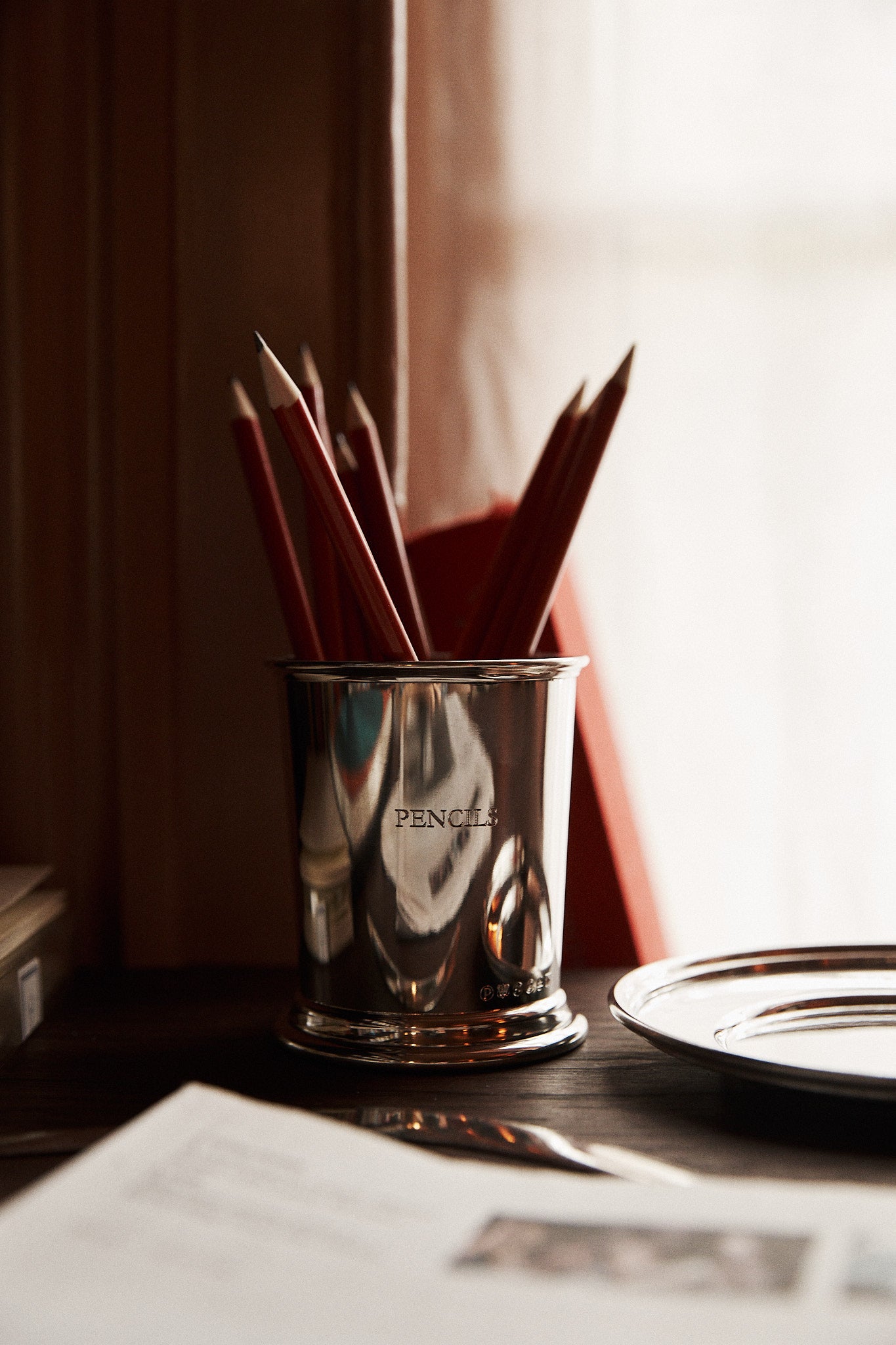 The Serena Set with Pencil Cup and Pencils features a pewter cup labeled “PENCILS” holding red pencils on a wooden desk beside a plate and open book, illuminated by soft daylight from a nearby window.
