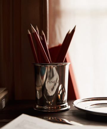 The Serena Set with Pencil Cup and Pencils features a pewter cup labeled “PENCILS” holding red pencils on a wooden desk beside a plate and open book, illuminated by soft daylight from a nearby window.