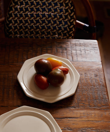 Four vibrant tomatoes sit on a Cirrus Octagonal Plate atop a wooden table, with a patterned chair in the background.