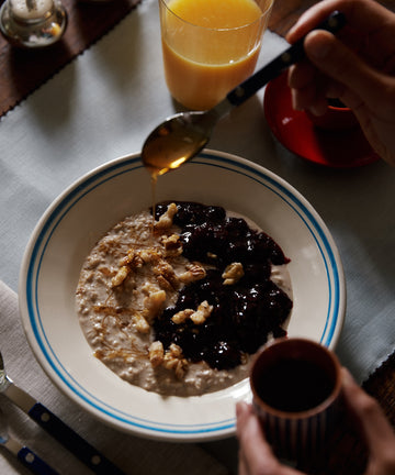 A bowl of oatmeal in the Folk Pasta Bowl, Jester, topped with walnuts and fruit compote, honey drizzle, orange juice, and mix-and-match tableware on the table.