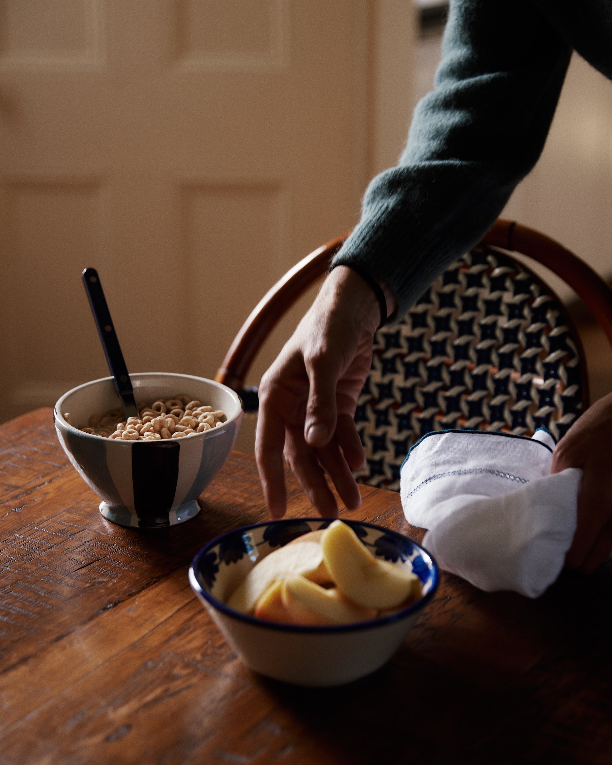 A hand reaches for sliced apples in the Mina Bowl, Set of 4, featuring a unique stripe design on a wooden table with cereal, a spoon, and a folded napkin.