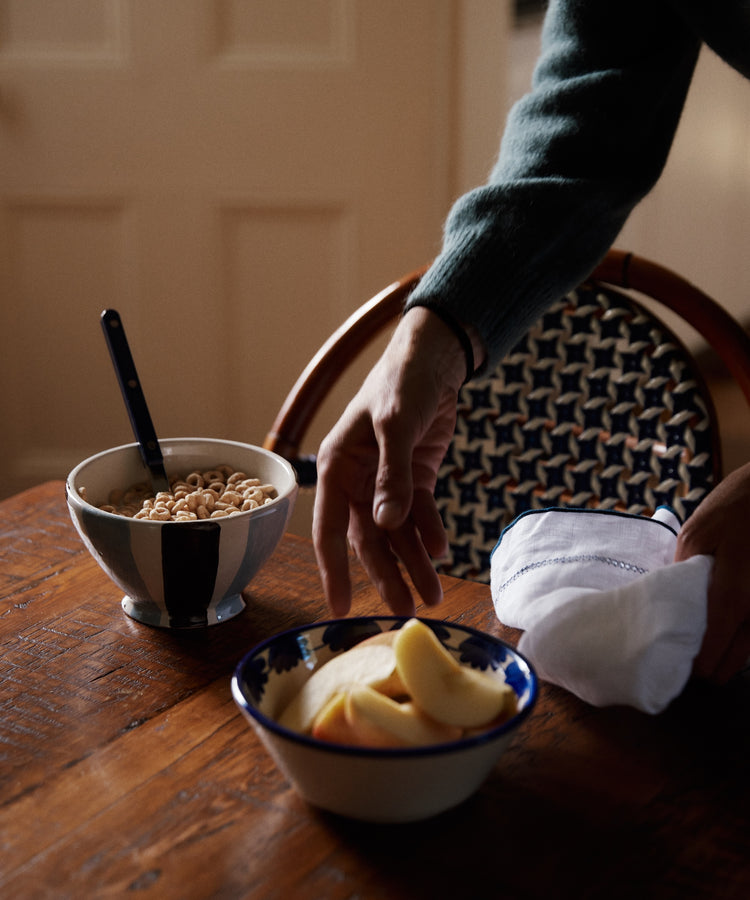 A hand reaches for sliced apples in the Mina Bowl, Set of 4, featuring a unique stripe design on a wooden table with cereal, a spoon, and a folded napkin.