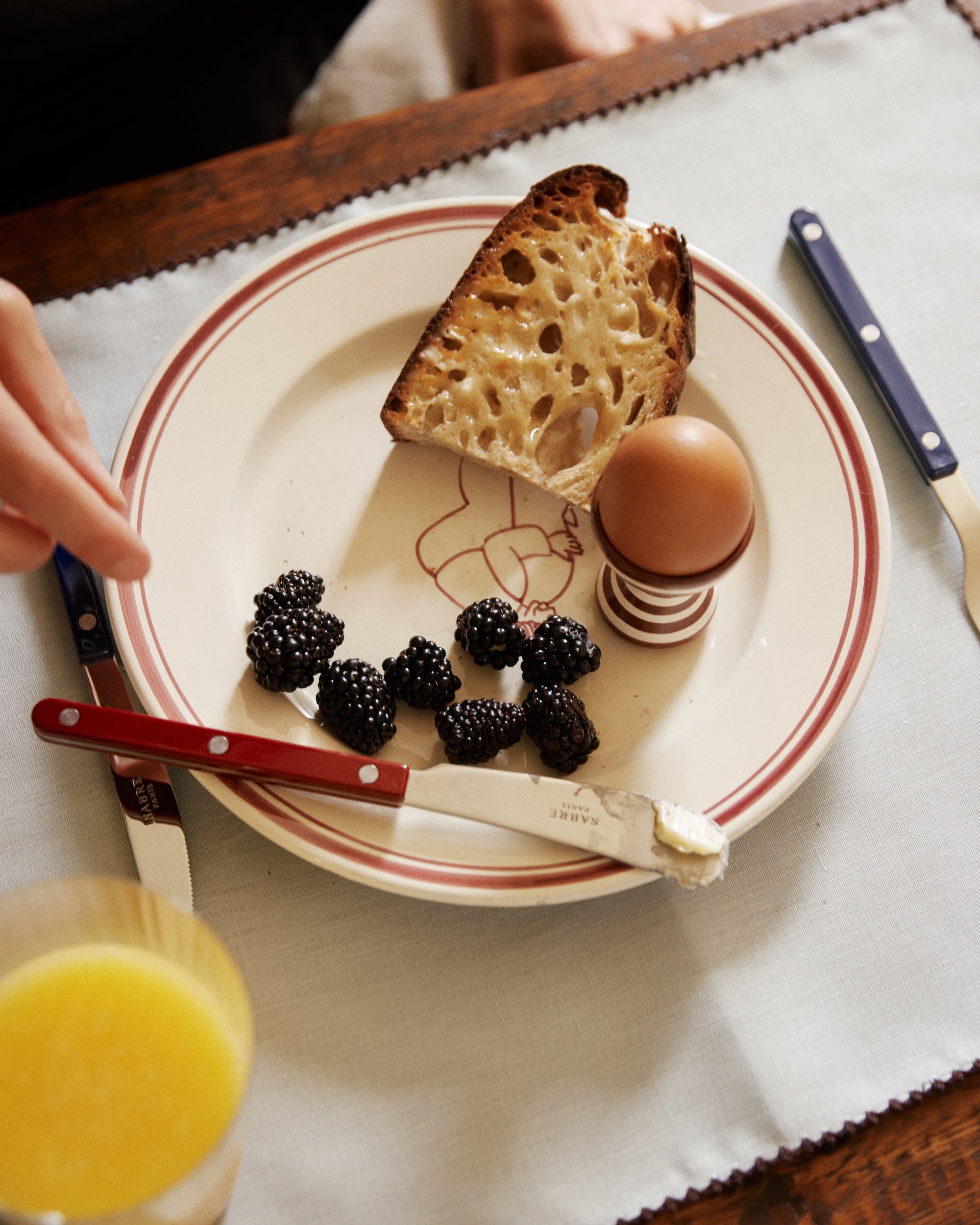 The Folk Pasta Bowl, Farmer holds bread, a hard-boiled egg in a holder, blackberries, a buttered knife, and a glass of orange juice.