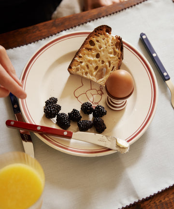 The Folk Pasta Bowl, Farmer holds bread, a hard-boiled egg in a holder, blackberries, a buttered knife, and a glass of orange juice.
