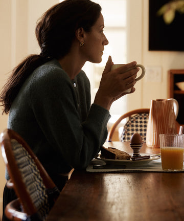 A woman sits at a table holding a mug, with the Natalia Egg Cup, Set of 2, toast, orange juice, and a pitcher in front of her.