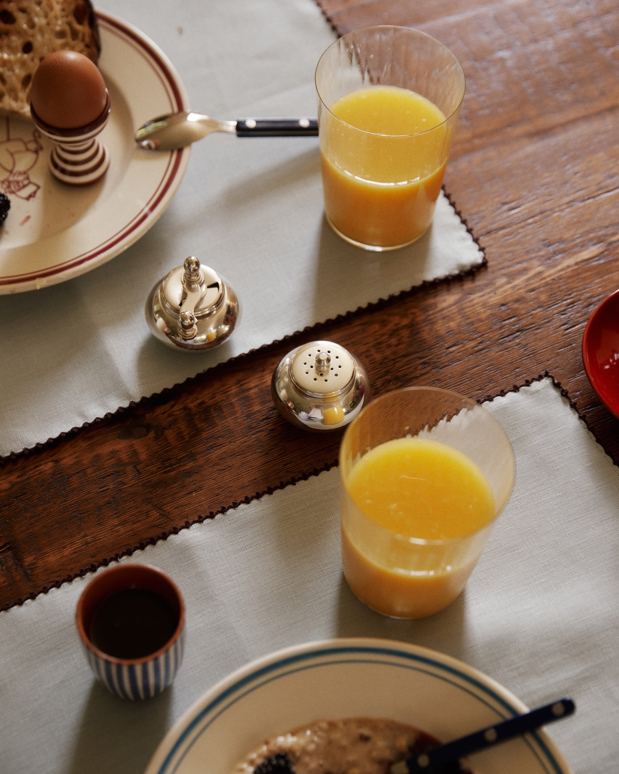 Two glasses of orange juice, a soft-boiled egg, bread, oatmeal, a small cup, and the Jules Pepper Mill and Salt Shaker on a wooden table.