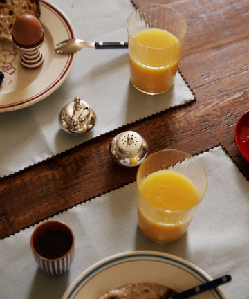 Two glasses of orange juice, a soft-boiled egg, bread, oatmeal, a small cup, and the Jules Pepper Mill and Salt Shaker on a wooden table.
