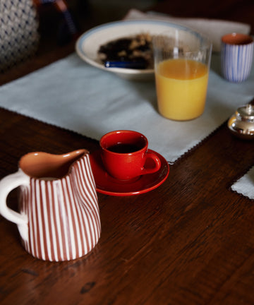 The Gio Espresso Set, a glass of orange juice, a striped ceramic pitcher, and a plate with food sit on a wooden table with blue placemats.