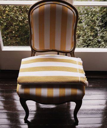A wooden chair with yellow and white striped upholstery sits by the window, topped with a neatly folded Citrino Stripe Towel—its vibrant cotton stripes echoing the chair’s pattern and brightening the sunlit corner.