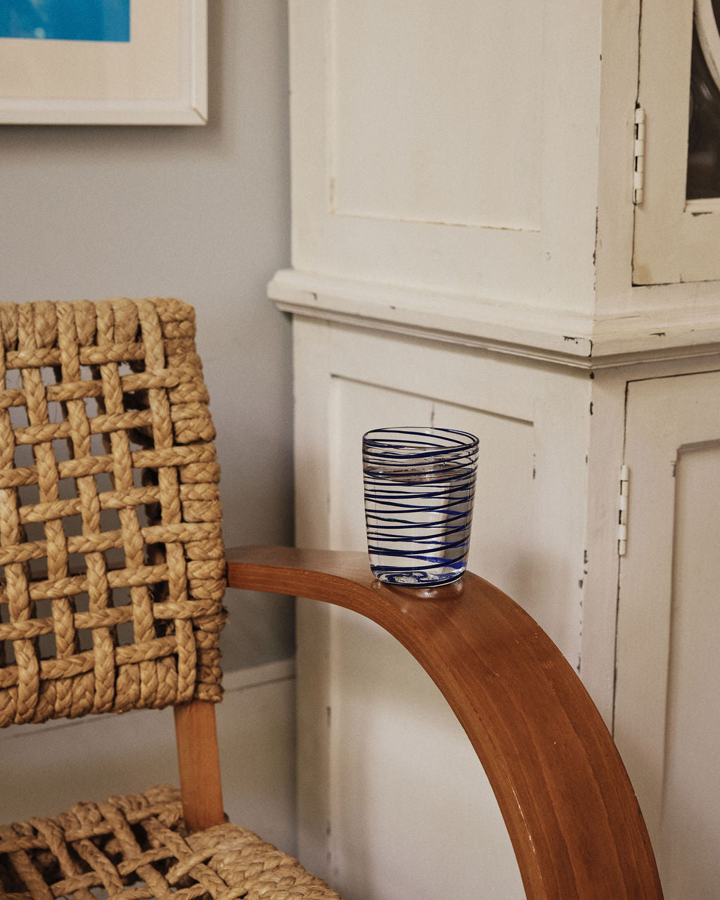 A woven chair with a wooden armrest holds a Spirale Glass, Set of 2—clear Murano glasses with blue stripes—placed beside a white cabinet against a beige wall.
