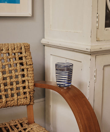 A woven chair with a wooden armrest holds a Spirale Glass, Set of 2—clear Murano glasses with blue stripes—placed beside a white cabinet against a beige wall.