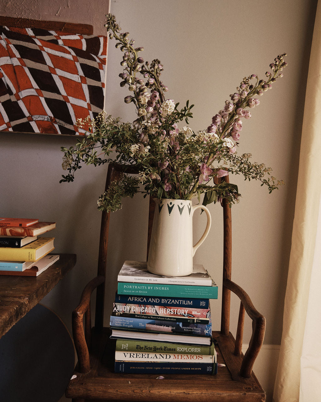 A Vito Jug with flowers rests on a wooden chair beside a stack of books, a table, and a painting—making this handmade piece a unique gift for any home.