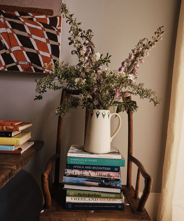 A Vito Jug with flowers rests on a wooden chair beside a stack of books, a table, and a painting—making this handmade piece a unique gift for any home.