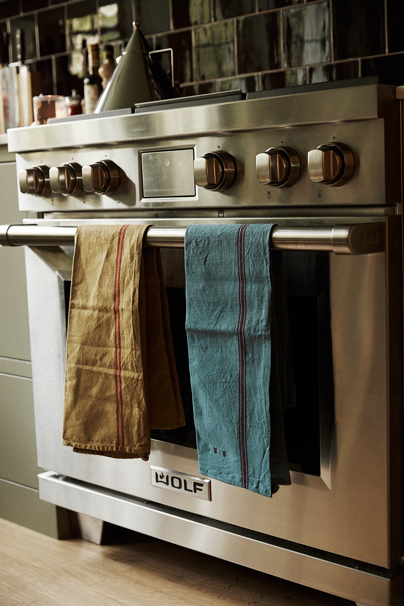 A stainless steel Wolf oven with French Vintage Tea Towels in brown and blue hanging on the handle; stove knobs and kitchen items are visible in the background, adding a touch of Provence charm to the kitchen.