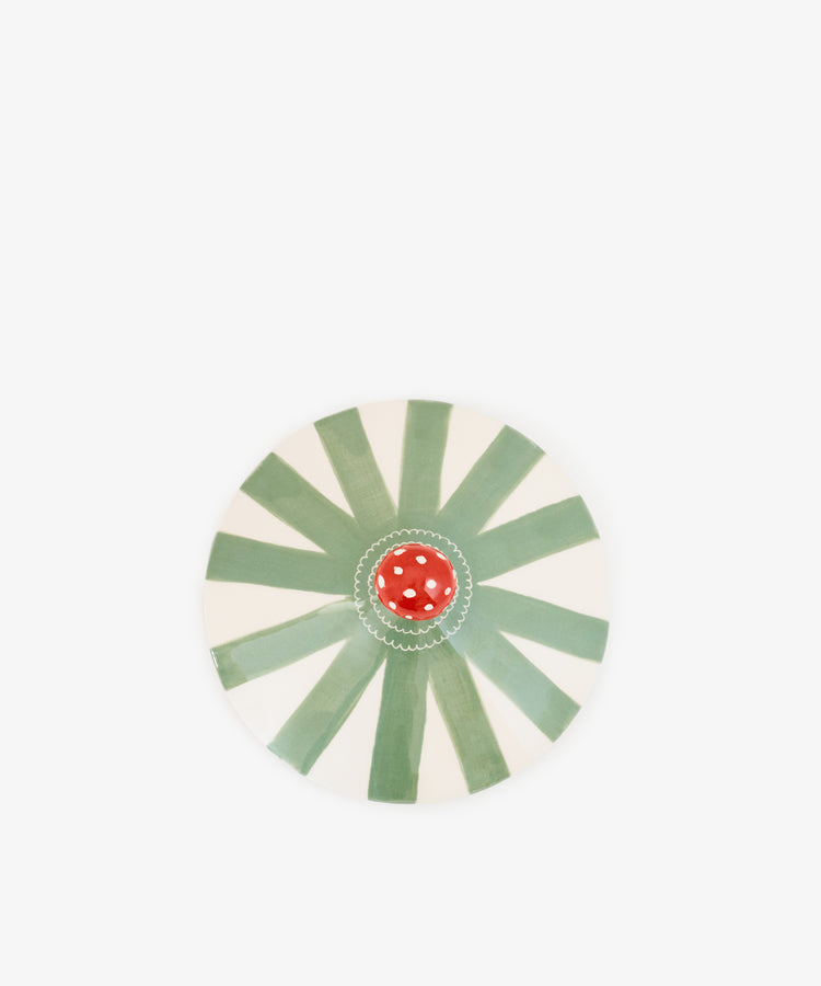 Hand-crafted in Portugal, the Magic Box is a round plate adorned with green and white radial stripes reminiscent of a circus tent and features a small red mushroom with white spots at its center, set against a plain white background.