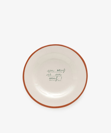 The Un oeuf is un oeuf plate is crafted from ceramic stoneware and features a brown rim with the phrase handwritten in green cursive at the center, reflecting Laetitia Rougets playful hand-painted style.