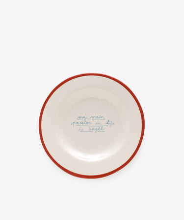 The My main passion in life is bagel plate by Laetitia Rouget is handmade, white with a red rim, and features blue handwritten text in the center.