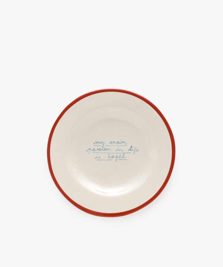 The My main passion in life is bagel plate by Laetitia Rouget is handmade, white with a red rim, and features blue handwritten text in the center.