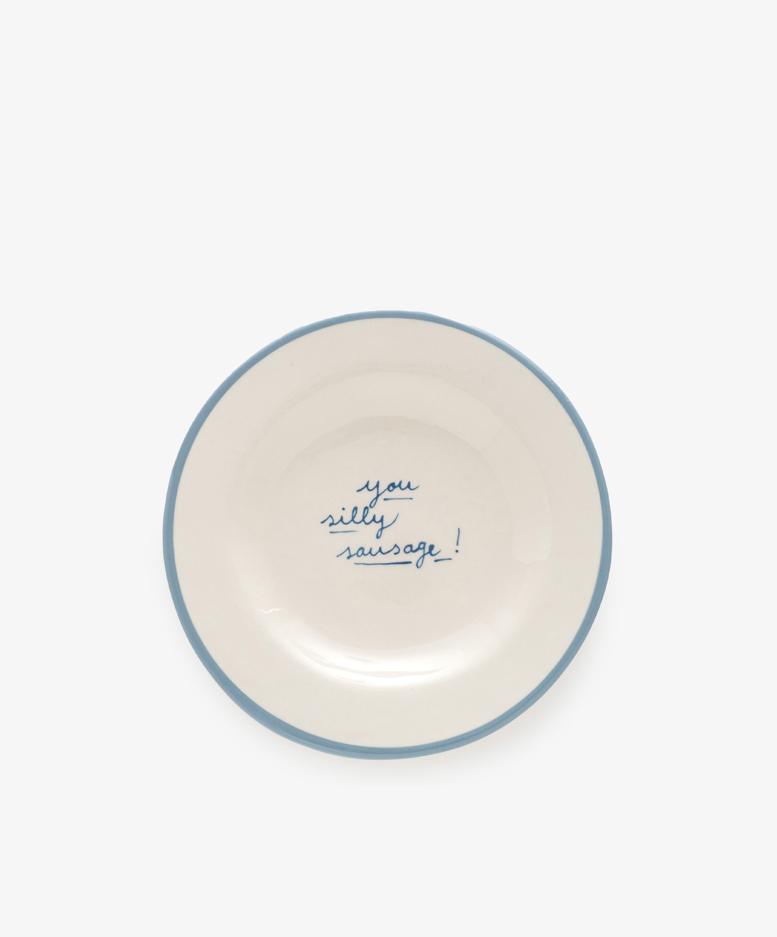 The You silly sausage! plate is a white ceramic dish, handmade in Portugal by artist Laetitia Rouget, featuring a blue rim and the phrase “you silly sausage!” handwritten in blue at the center.