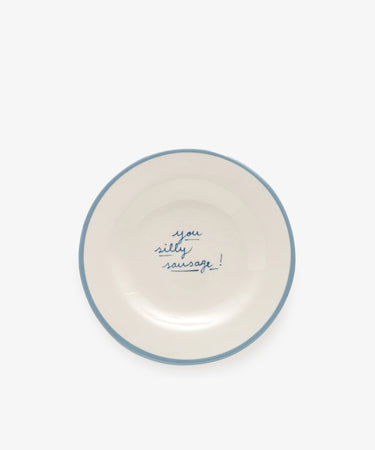The You silly sausage! plate is a white ceramic dish, handmade in Portugal by artist Laetitia Rouget, featuring a blue rim and the phrase “you silly sausage!” handwritten in blue at the center.