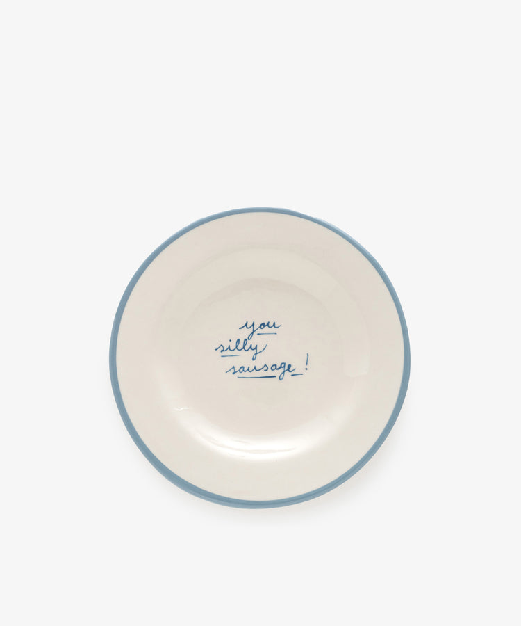 The You silly sausage! plate is a white ceramic dish, handmade in Portugal by artist Laetitia Rouget, featuring a blue rim and the phrase “you silly sausage!” handwritten in blue at the center.