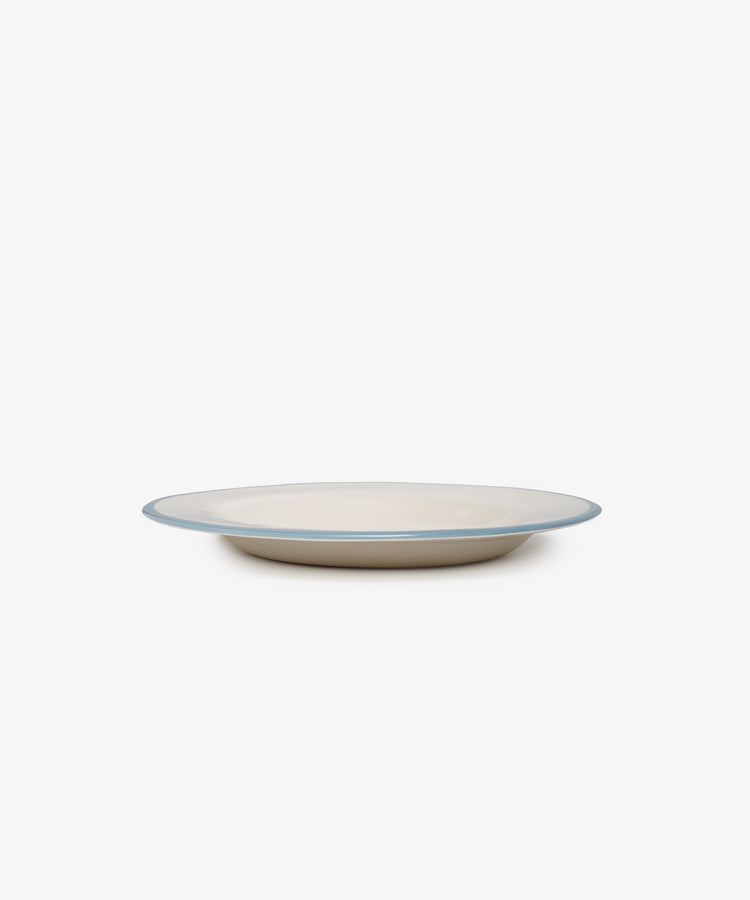 A plain, round, beige ceramic plate named You silly sausage! with a thin blue rim—crafted in Laetitia Rouget’s style—is shown from the side against a white background.